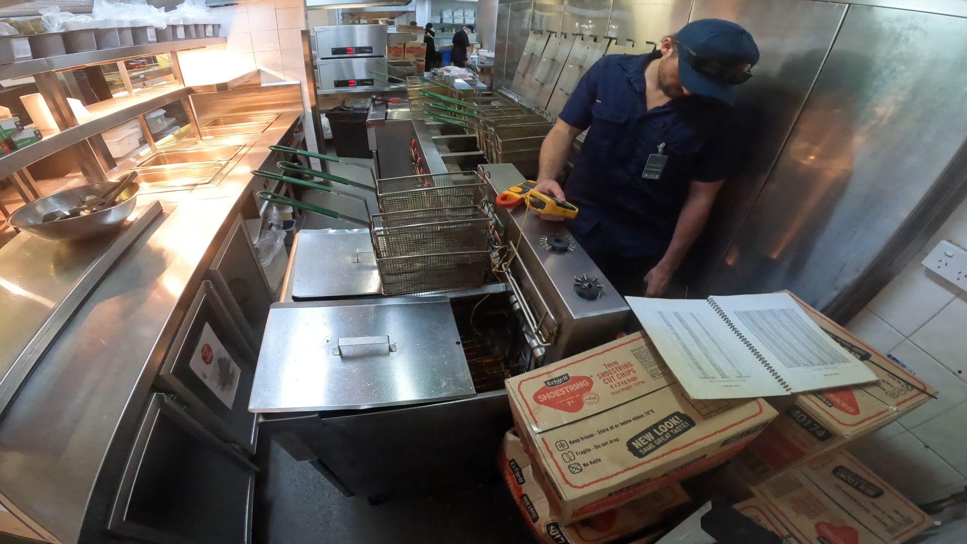 CHEFTECH technician testing a commercial deep fryer thermostat with a digital meter in a busy kitchen